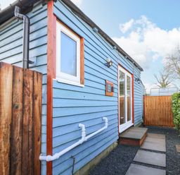 An exterior view of a blue wooden building with a white window and door surrounded by a wooden fence and green hedge at Shed Your Blues Brilley near Kington