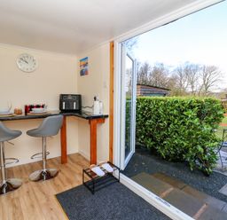 A small dining area with two stools and a countertop with plates and glasses next to an open door leading to a garden with bushes and a metal chair at Shed Your Blues in Brilley near Kington