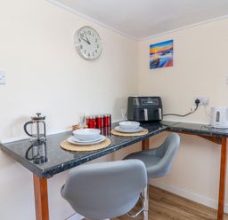 A kitchen corner with a black countertop table set for two with plates and glasses a clock on the wall and kitchen appliances at Shed Your Blues in Brilley near Kington