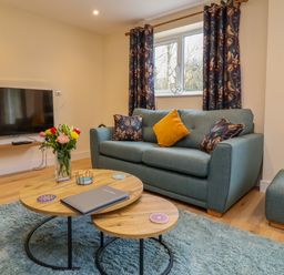 A living room with a teal sofa and matching ottoman a wooden coffee table with flowers and coasters mounted TV and floral curtains at Hideaway in Great Torrington