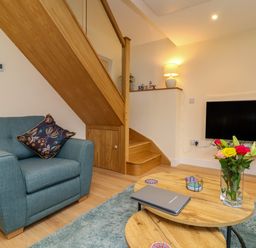 A living room with a blue armchair wooden staircase a wall-mounted television a lamp a wooden coffee table with flowers and a laptop at Hideaway Great Torrington