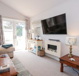 A living room with a beige sofa wooden tables a wall mounted television and glass doors at Ocean Breeze in Abersoch