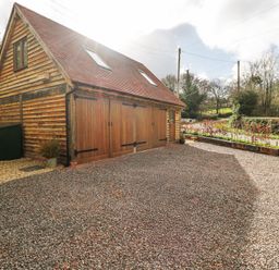 A wooden building with large wooden doors and a gravel driveway surrounded by trees and shrubs at Pheasants Rise Apartment in Ross-On-Wye