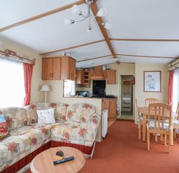 A living room with a floral patterned sofa a wooden coffee table a dining table with four chairs and a kitchen area at 120 Caravan in Hemsby
