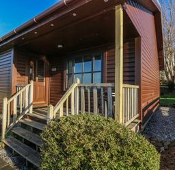 The exterior of a brown wooden cabin with a small porch and steps surrounded by bushes and grass at Creag Mhor Lodge in Back of Keppoch near Arisaig