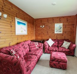 A living room with two red patterned sofas a matching ottoman wooden paneled walls and framed pictures at Creag Mhor Lodge Back of Keppoch near Arisaig