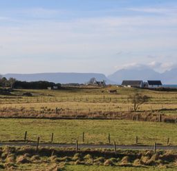 A rural landscape with fenced fields, houses, cows, a road, and mountains in the background at Glen Mhor Lodge in Arisaig