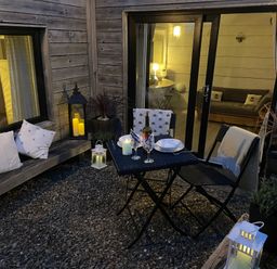An outdoor patio area with a small table set for dining two with wine and candles and a bench with cushions at The Cow Shed in Porthcothan near Mawgan Porth