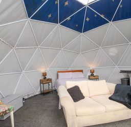An interior of a dome with a bed two bedside tables with lamps a beige sofa with cushions a floral chair and a kitchenette at Meadow View - Bryn-Mawr Retreat in Brynmawr near Llanymynech