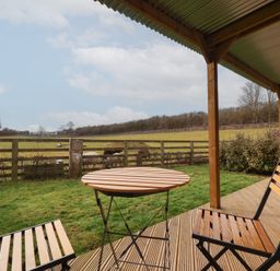 A wooden table and two chairs on a covered deck overlooking a grassy field with a wooden fence and sheep at Beech Tree Cabin in Goodmanham near Market Weighton
