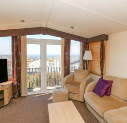 A living room with beige sofa and armchair next to glass doors with curtains and a television on a wooden stand at 21 Bay View in Maenygroes near New Quay Ceredigion