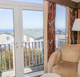 A beige armchair next to a floor lamp by windows with floral curtains and blinds overlooking a balcony with sea view at 21 Bay View in Maenygroes near New Quay Ceredigion