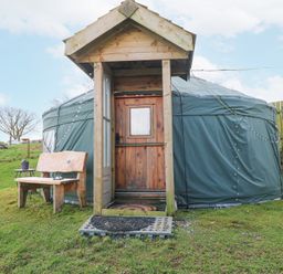 A green yurt with a wooden door and porch with a bench and small table on grass at Blaidd Bach in Carmarthen