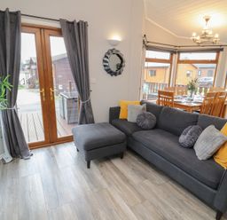 A living room with a gray sofa and ottoman near wooden doors and a dining area with wooden chairs and a table at Lakeside 18 in Carnforth