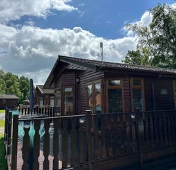 A wooden cabin with a fenced deck and outdoor furniture under a partly cloudy sky at Lakeside 18 in Carnforth