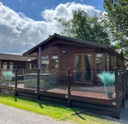 A wooden cabin with a deck and glass railing surrounded by grass and trees at Lakeside 18 in Carnforth