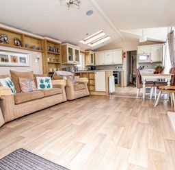 An open plan living area with a beige sofa and armchair a dining table with chairs a kitchen with cabinets and appliances and wooden flooring at Hazel Park Lodge HP4 Rhyl