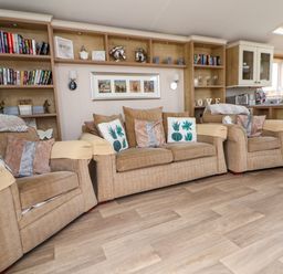 A living room with three beige armchairs, cushions with floral patterns, built-in bookshelves with books and decorative items, and a kitchen area in the background at Hazel Park Lodge HP4 in Rhyl