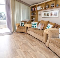 A living room with beige sofas and armchairs with patterned cushions and shelves with decorations at Hazel Park Lodge HP4 in Rhyl
