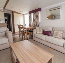 A living room with beige sofas, a wooden coffee table, dining table with chairs near a window and patterned curtains at Hazel Park 11 HP11 in Rhyl