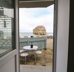 View through an open door showing a small outdoor area with a white metal table and three chairs overlooking the sea and a rock formation at The Beach Apartment in South Shields