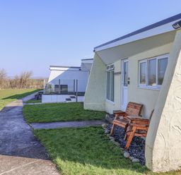 Outdoor area with a concrete walkway grass patches and a small patio with two wooden chairs at Atlantic Escape in Widemouth Bay