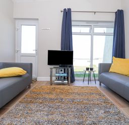A living room with two gray sofas with yellow cushions a television on a glass stand a small round table with two mugs and blue curtains at Atlantic Escape in Widemouth Bay