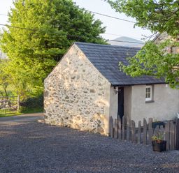 A small stone and plaster building with a slate roof next to a fenced patio with white metal chairs and tables at Cwt Mochyn in Llanberis