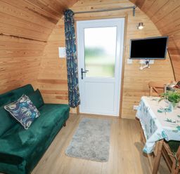 A small wooden room with a green sofa a floral pillow a door a wall-mounted tv a table with a floral tablecloth and chairs with a plant and a wine bucket Chantelles Holbeach