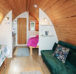 A small cabin interior with wooden walls featuring a green sofa with a patterned pillow a dining table with a floral tablecloth a bed with a pink blanket and a wooden door in the back at Chantelles in Holbeach