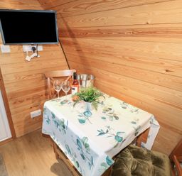 A small dining area with a table covered by a floral tablecloth two wooden chairs a bottle of wine two wine glasses a metal ice bucket a small plant a wall-mounted flat screen television and wooden panel walls at Chantelles in Holbeach