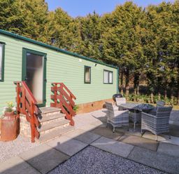 An outdoor patio area with wicker chairs and a round table next to a green mobile home with wooden steps and trees in the background at Lodge 1 in Silverdale
