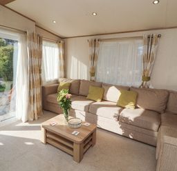 A sitting room with a beige corner sofa green cushions wooden coffee table with flowers and large windows with curtains at Lodge 1 in Silverdale