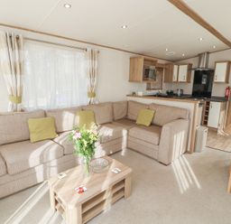 A living room with a beige sectional sofa and green cushions a wooden coffee table with flowers and coasters a kitchen area with cabinets and appliances in the background at Lodge 1 in Silverdale