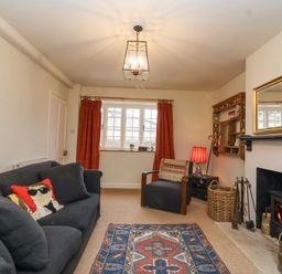 A living room with a black sofa with decorative pillows an armchair a patterned rug a fireplace with a wood-burning stove and a window with red curtains at Little Lemhill Farm Cottage in Lechlade-On-Thames