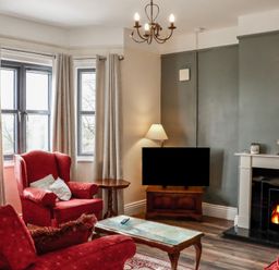 A living room with red armchairs a fireplace with fire a television on a wooden cabinet and windows with curtains at Heatherbell House Ballyhoura near Buttevant