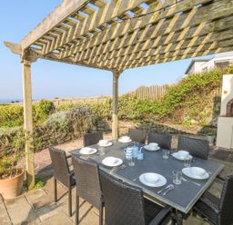 An outdoor dining area with a table and chairs under a wooden pergola with a built in brick barbecue and garden in the background at Coastmans Nest in Mawgan Porth