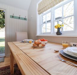 A wooden dining table with plates cups toast and a bowl of tomatoes next to a window with flowers at The Woodshed in Tendring near Weeley
