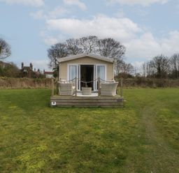 A small beige cabin with a wooden deck and outdoor chairs on a grassy lawn at Breathing Space in Happisburgh near Walcott