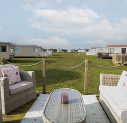 An outdoor seating area with wicker furniture and cushions overlooking a grassy area with caravans at Breathing Space Happisburgh near Walcott