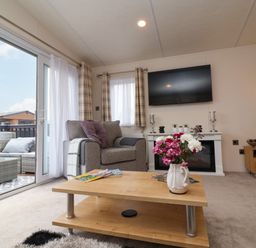 A living room with a gray armchair wooden coffee table with flowers and magazines and a wall-mounted TV at Lodge 36 - Kingfisher Lodge in Wickhamford near Evesham