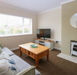 A living room with a sofa with cushions a wooden coffee table a flat screen TV on a cabinet a fireplace and a round mirror at Little Lodge in Dersingham