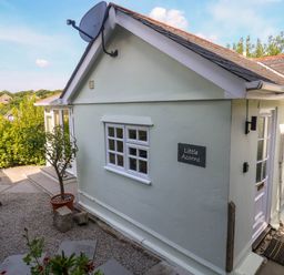 A small exterior side view of a light green building with a window a door a satellite dish and a sign that says Little Acorns with outdoor seating and plants at Little Acorns in Gweek