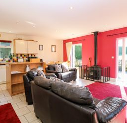An open living area with black leather sofas a wood stove with protective screen red walls and an adjacent kitchen with tiled backsplash and white cabinets at Swallow Barn in Holton near Halesworth