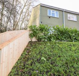 An exterior view of a green house with white windows behind a hedge and wooden railing on a grassy slope at Arnside House in South Lakeland Leisure Village