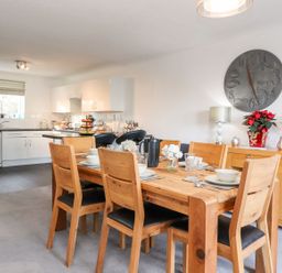 A dining area with a wooden table set with dishes and glasses next to a kitchen with white cabinets and black appliances at Arnside House in South Lakeland Leisure Village