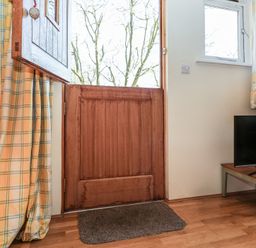 An open wooden stable door with a window and curtains next to a television on a wooden floor at Woodland Retreat Lodge in Brundish near Laxfield