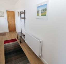 A narrow hallway with a wooden door a multi-colored rug a coat rack with a bench a radiator and framed pictures on white walls at Arfryn in Solva