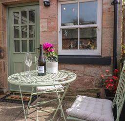 A small outdoor seating area with a metal table and chairs with cushions flowers on the table and against the house at Dromore Cottage in Norham