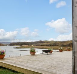 A wooden deck with flower pots and a fire pit overlooking a water body and hills at WildLuing in Cullipool Isle Of Luing